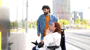Modern bir tramvay durağında tekerlekli sandalyeli bir kadına yardım eden, programı kontrol eden ve destek öneren genç bir adam.