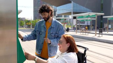Tekerlekli sandalyedeki bir kadına yardım eden nazik genç bir adam açık hava istasyonunda tren biletini onaylıyor.