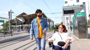 Şehir hareketliliğini araştıran neşeli bir çift, tekerlekli sandalye kullanan bir kadın, tramvay durağında birlikte yürüyen, halka açık alandaki sıcaklık ve bağlantıyı paylaşan bir kadın.