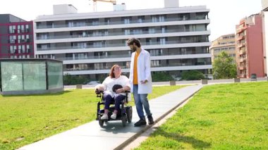 Motorlu tekerlekli sandalyedeki kadın hastaya yardım eden dost canlısı doktor güneşli bir şehir parkında geziniyor.