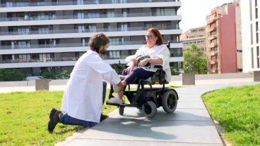 Şefkatli tıbbi uzmanlar elektrikli sandalye ayaklığı ayarlıyor, açık hava parkından hoşlanan engelli kadınlara kişisel destek sağlıyor.