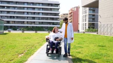 Nazik doktor tekerlekli sandalyeli neşeli engelli bir kadınla yürüyor, güneşli bir günün tadını çıkarıyor.