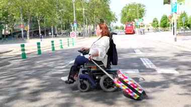 Tekerlekli sandalye kullanan beyaz kadın, yanında renkli valizlerle toplu taşıma durağında sabırla bekliyor, trafik akışını izliyor.