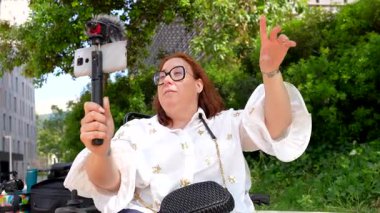 Tekerlekli sandalyedeki tutkulu kadın, çevrimiçi içerik yaratmak için dış mikrofon ve sabitleyici kullanan akıllı telefon kullanıyor.