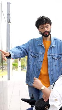 Genç bir adam, elektrikli sandalyedeki bir kadına şehir caddesinde yol göstererek yardım ediyor.