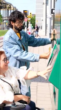 Tekerlekli sandalyedeki gülümseyen engelli bir kadın için bilet parası ödeyen kibar bir adam.