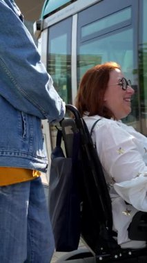Destekleyici bakıcı şehir caddesinde geziniyor, tekerlekli sandalyedeki gülümseyen engelli kadını tramvay durağının yakınına itiyor, hareketli, bağımsız ve neşeli.