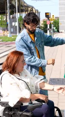 Tekerlekli sandalyedeki kadına yardım eden, şehir tramvayı biletini onaylayan, ulaşılabilirliğini ve toplum desteğini gösteren adam.