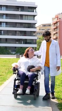 Dost canlısı doktor tekerlekli sandalyedeki mutlu engelli bir kadına yardım ediyor güneşli bir şehir parkında geziniyor.