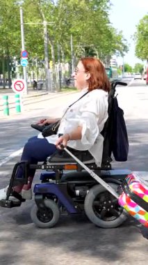 Elektrikli sandalye kullanan kadın gezgin, renkli bavulla güvenli bir şekilde şehir tramvay durağında bekliyor, güneşli bir günde ulaşılabilir ve bağımsız hareketliliği somutlaştırıyor.