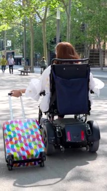 Kadın gezgin şehir parkında geziniyor, elektrikli sandalye kullanırken renkli bavul çekiyor, aktif yaşam tarzı için bağımsızlığı ve hareketliliği somutlaştırıyor
