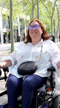 Engelli neşeli kadın tatilinin tadını çıkarıyor, elektrikli tekerlekli sandalyesiyle şehir caddesinde renkli bir bavul taşıyor.