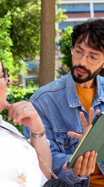 Destekleyici genç adam tekerlekli sandalye kullanıcısıyla dijital bilgi paylaşıyor, yeşil şehir parkı ortamında kapsayıcı iletişim gösteriyor