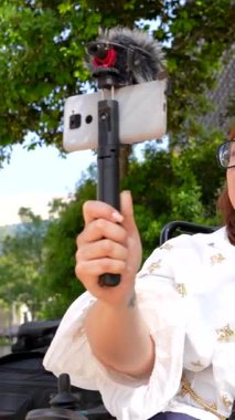 Tekerlekli sandalyedeki tutkulu içerik yaratıcısı dışarıda bir video blogu kaydediyor. Harici mikrofonu olan bir akıllı telefon kullanıyor.
