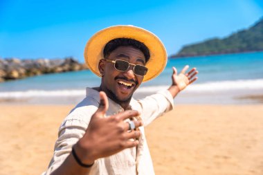 Şık bir şapka ve son moda güneş gözlüğü takan genç adam, güzel bir tropikal plajda kaygısız bir yaz tatilinin tadını çıkarıyor.