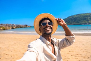 Genç bir adam tropik bir plajda selfie çekiyor, yaz tatilinin tadını çıkarıyor ve güneş gözlüğü ve hasır şapka takıyor.