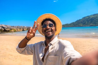 Şapkalı ve güneş gözlüklü genç adam tropik bir plajda selfie çekerken zafer işareti yapıyor.