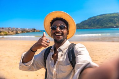Sırt çantası ve hasır şapkalı genç siyahi adam güneşli bir sahilde selfie çekerken başparmağını kaldırıyor.
