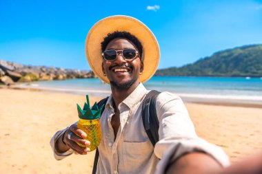 Hasır şapkalı ve güneş gözlüklü genç siyah adam tropik bir plajda ferahlatıcı bir içki içip selfie çekiyor.