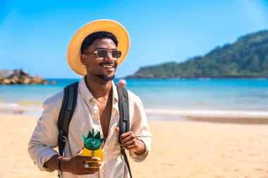 Genç siyah adam tropik bir sahilde yürüyor, ananas şeklinde bir kokteyl tutuyor ve sırt çantası, hasır şapka ve güneş gözlüğü takıyor.