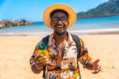 Genç adam, güneşli bir sahilde ferahlatıcı bir tropikal içecek tutarken gülümsüyor ve yaz tatili kaçamağının tadını çıkarıyor.
