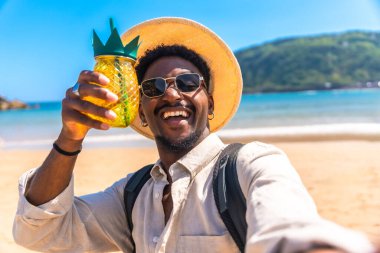 Güneş gözlüğü ve hasır şapka takan genç adam sahilde selfie çekerken ananas şeklinde kokteyl tutuyor.
