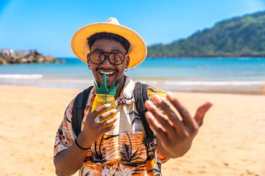 Genç siyah bir adam güneşli bir sahilde ferahlatıcı bir kokteylin tadını çıkarıyor, canlı atmosferin ve kaygısız yaz havasının tadını çıkarıyor.