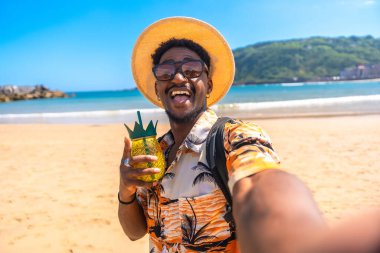 Genç adam güneşli bir sahilde tropikal bir kokteylin tadını çıkarıyor. Tatilin tadını çıkarırken selfie çekiyor.