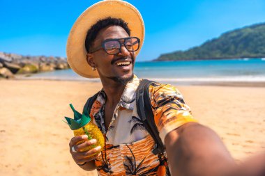 Genç siyahi adam yaz tatilinin tadını çıkarıyor. Tropikal bir plajda ananas şeklinde bir kokteyl tutarken selfie çekiyor.