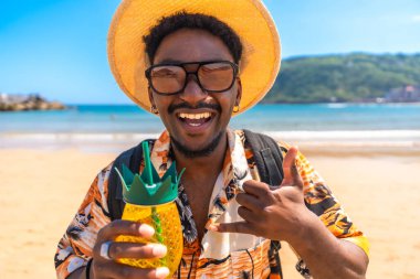 Genç adam tropik bir plajda yaz tatilinin tadını çıkarıyor, ananas şeklinde bir kokteyl tutuyor ve shaka işareti yapıyor.