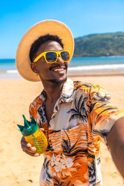 Güneş gözlüğü ve hasır şapka takan genç adam güneşli bir sahilde selfie çekerken tropikal bir içkinin tadını çıkarıyor.