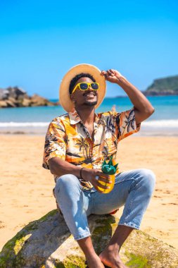 Güneş gözlüğü ve hasır şapka takan genç adam güneşli bir plajda bir kayanın üzerinde dinlenirken ananas şeklinde bir kokteyl tutuyor.