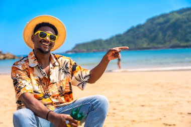 Hawai gömleği, güneş gözlüğü ve hasır şapka takan siyahi bir adam tropikal bir plajda bir şeyi işaret ediyor.