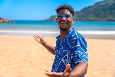 Güneş gözlüklü ve Hawaii gömlekli genç siyahi adam yaz tatilinin tadını çıkarıyor. Açık kollarıyla tropik bir plaja gelen insanları selamlıyor.