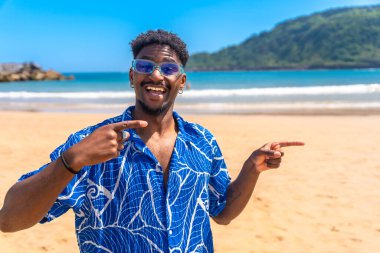 Genç adam güneşli bir sahilde gülümserken işaret parmaklarıyla kendini ve yan tarafı işaret ediyor.
