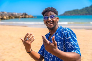 Genç adam tropikal bir plajda yaz tatilinin tadını çıkarıyor. Geniş bir şekilde gülümsüyor ve neşeyle el kol geziyor. Güneş ve okyanus esintisini emiyor.