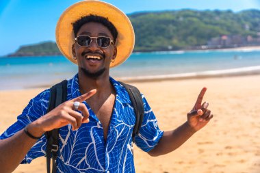 Mutlu siyah turistler gülümserken işaret parmaklarını güzel bir sahile doğrultup güneşin ve denizin tadını çıkarıyorlar.