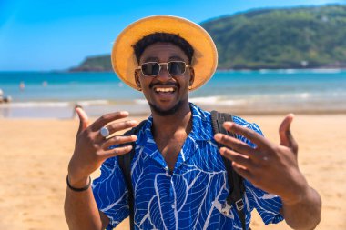 Şapka takan ve güneş gözlüğü takan genç adam güneşli tropik plaj ve okyanus manzarasının tadını çıkarırken sevinçle el kol hareketi yapıyor.