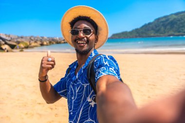 Hasır şapkalı ve güneş gözlüklü genç siyahi adam güneşli bir sahilde selfie çekerken başparmağını kaldırıyor.