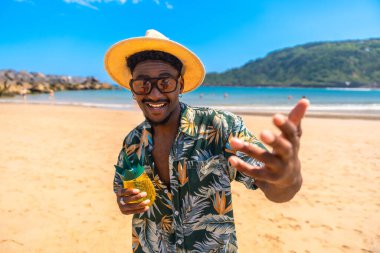 Hawaii gömleği ve güneş gözlüğü takan siyah bir adam yaz tatilinin tadını çıkarıyor. Tropik bir sahilde kokteyl içiyor.