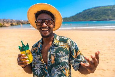 Hasır şapkalı, Hawaii gömlekli, gülümseyen ve güneşli bir sahilde ananas kokteyli tutan genç bir adam.