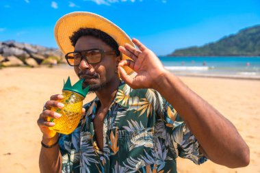Güneş gözlüğü takan ve hasır şapkalı genç adam güneşli bir sahilde ananas kokteyli içiyor.