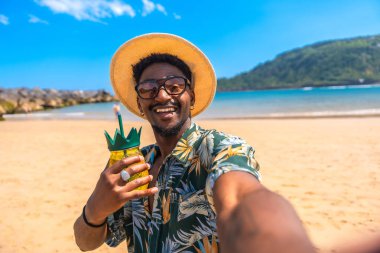 Genç adam güneşli bir sahilde güneşleniyor, ferahlatıcı bir kokteyl yudumluyor ve neşeli bir selfie ile neşeli anları yakalıyor.