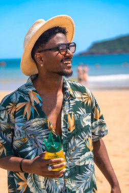Güneş gözlüğü ve şapka takan genç adam, ananas şeklinde bir içecek tutuyor, tropik bir sahilde yaz güneşinin tadını çıkarıyor.
