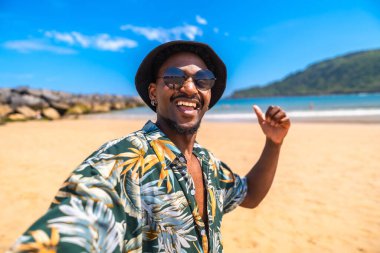 Genç adam tropikal bir plajda yaz tatilinin tadını çıkarıyor. Gülümserken selfie çekiyor, başparmak işareti yapıyor, mutluluk saçıyor.