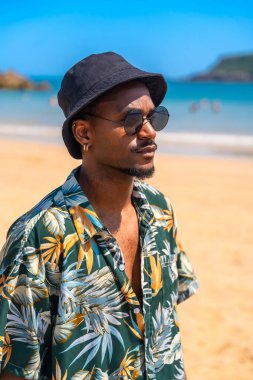 Güneş gözlüğü takan genç bir adam, Hawaii tişörtü ve kova şapkalı. Tropik bir plajda yaz tatilinin tadını çıkarıyor.