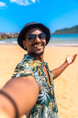 Genç adam, tropikal bir plajda selfie çekerken gülümsüyor, güneşin, kumun ve yaz tatilinin canlı atmosferinin tadını çıkarıyor.