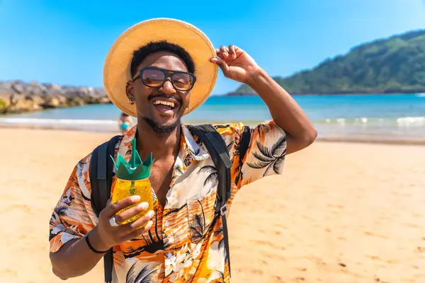 Hasır şapka ve güneş gözlüğü takan genç siyah adam güneşli bir sahilde dinlenirken ferahlatıcı bir ananas kokteylinin tadını çıkarıyor.
