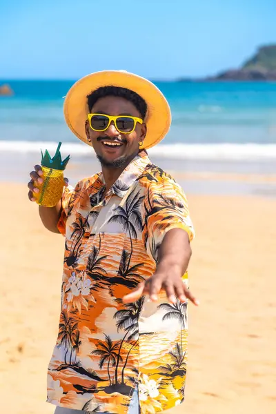 Hawaii gömleği ve güneş gözlüğü takan genç adam güneşli bir sahilde ananas kokteylinin tadını çıkarıyor.