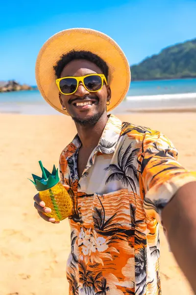 Genç bir adam tropikal bir plajda güneş gözlüğü ve hasır şapka takarak ananas şeklinde bir kokteyl tutuyor ve gülümsüyor.
